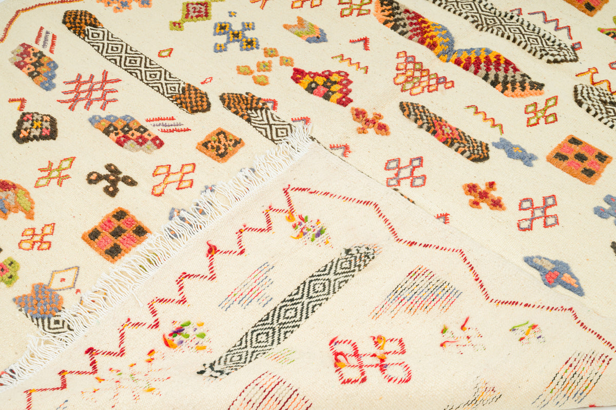 A close-up of a rug with an off-white base and colorful geometric motifs, including zigzags, diamonds, and abstract shapes in red, orange, yellow, blue, and black.