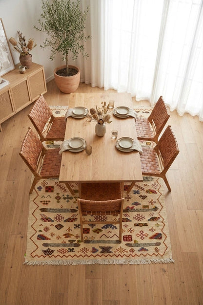 A styled dining room scene with a rectangular wooden table, four woven-back chairs, a patterned area rug, and a potted tree near sheer curtains and a side cabinet.