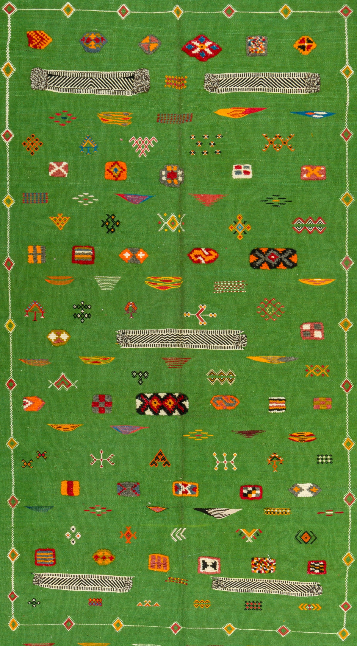 A green Moroccan rug featuring a dense pattern of colorful geometric motifs, including diamonds, crosses, and abstract shapes, bordered by a decorative patterned edge.