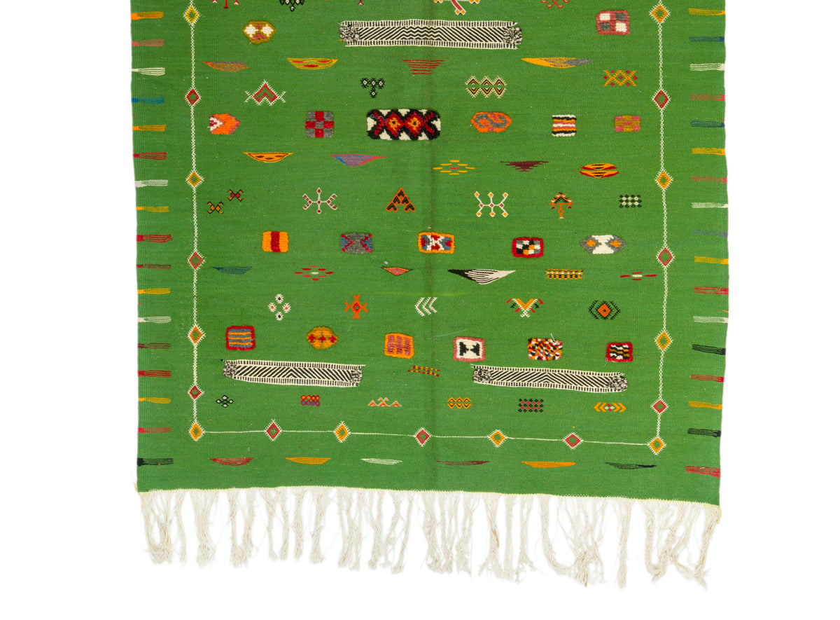 A green woven textile with white fringe at the bottom and an array of colorful geometric and symbolic motifs throughout, displayed flat against a white background.