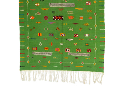 A green woven textile with white fringe at the bottom and an array of colorful geometric and symbolic motifs throughout, displayed flat against a white background.