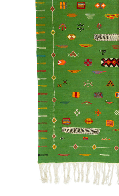 A close-up of a green moroccan rug with multicolored geometric motifs and white fringe along one end, shown against a white background.