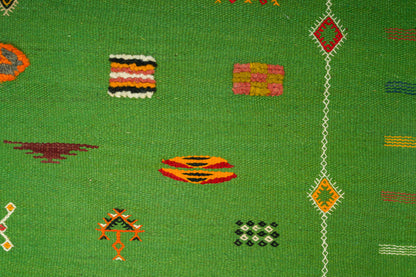 A close-up of a green Moroccan rug with scattered geometric and striped motifs in orange, red, yellow, pink, black, and white.