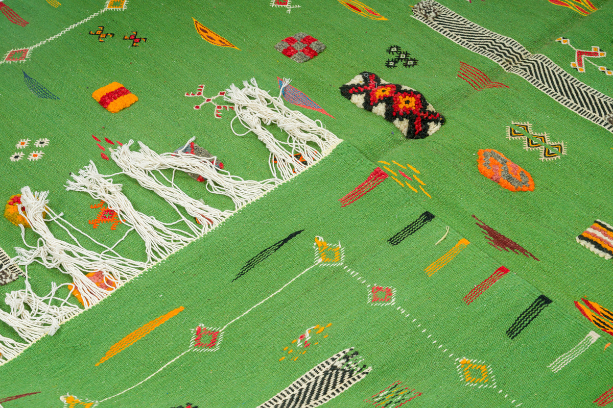 A close-up of a green  moroccan flat weave rug with colourful geometric motifs, white fringe, and a small multicolored woven accent on a matching green background.