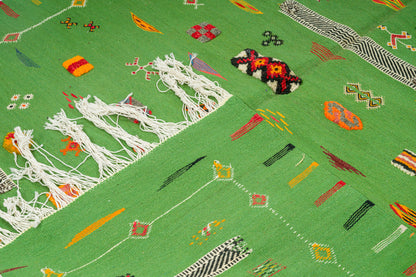 A close-up of a green  moroccan flat weave rug with colourful geometric motifs, white fringe, and a small multicolored woven accent on a matching green background.