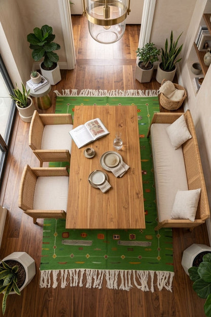Overhead view of a cozy dining nook with a rectangular wooden table, four woven chairs, a cushioned bench, and a green patterned rug on a wood floor, surrounded by potted plants.