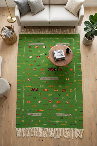 A green rectangular area rug with cream fringe and multicolored geometric patterns, styled in a modern living room setting on a light wood floor.