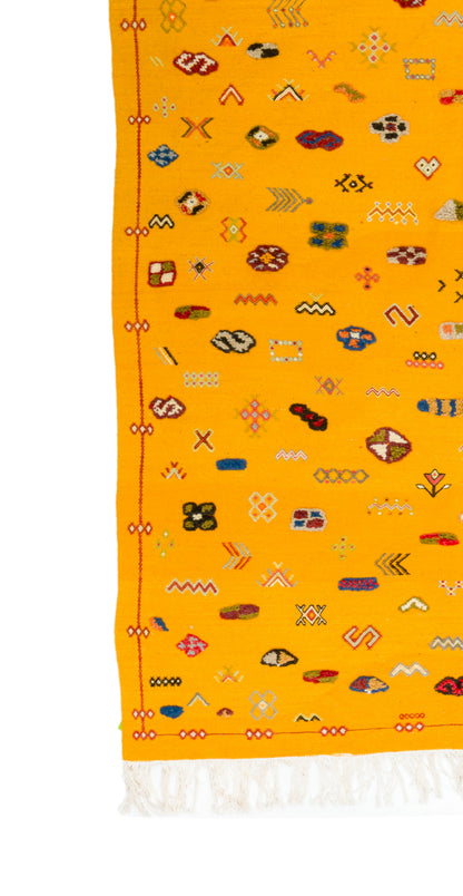 A close-up of a bright yellow rug with a white fringed edge and an allover scattered pattern of small multicolored geometric and abstract motifs.