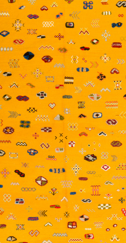 A close-up of a bright yellow rug or fabric with an all-over repeating pattern of small geometric and abstract motifs in red, blue, black, white, and brown.