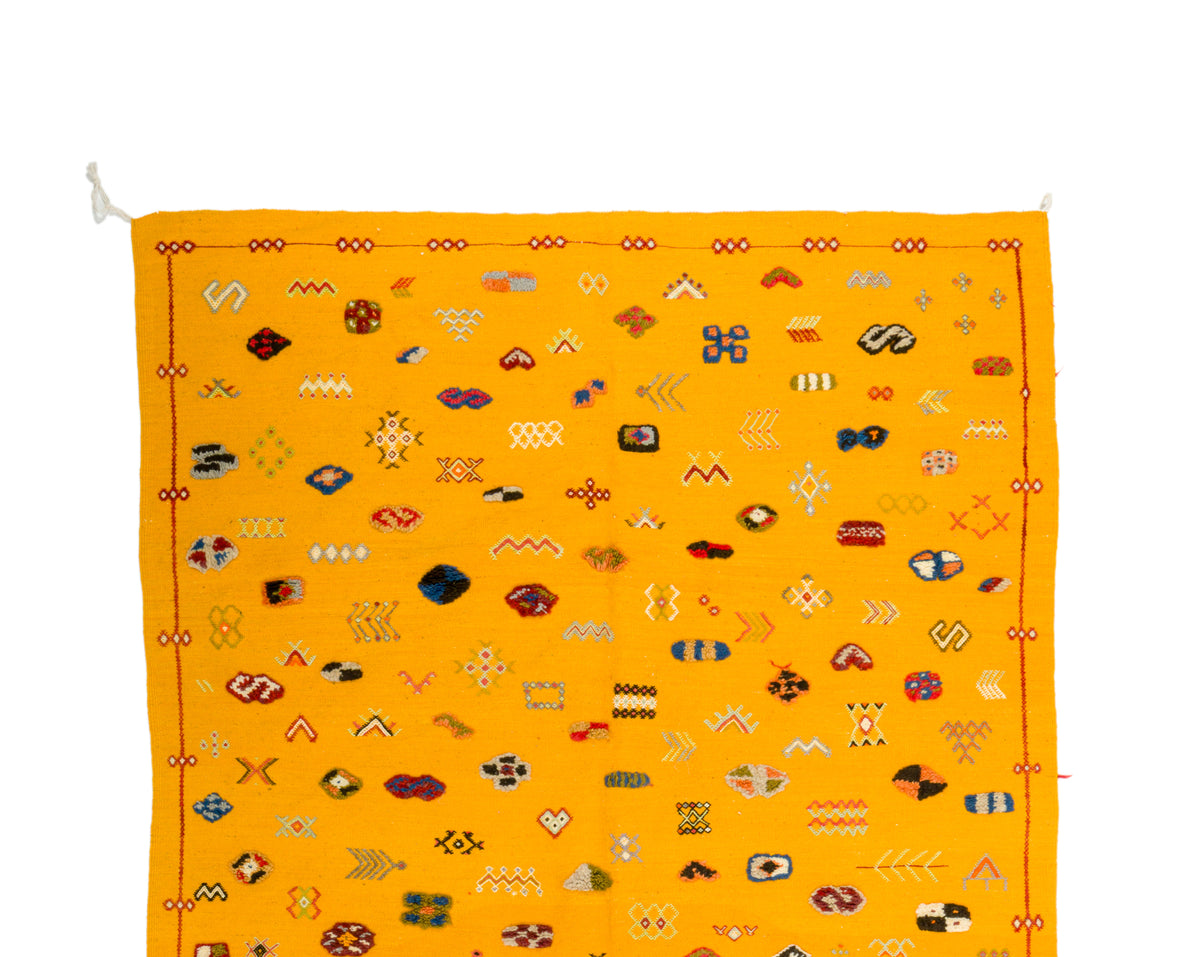A yellow rug with an all-over pattern of small multicolored geometric and symbolic motifs, bordered by red and orange decorative lines, shown flat against a white background.