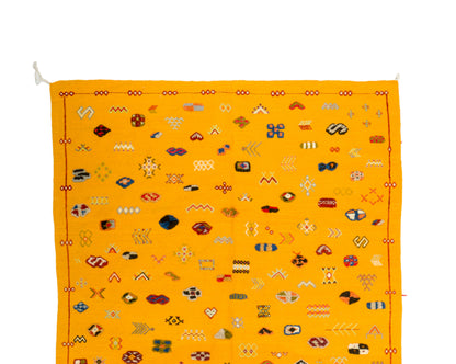 A yellow rug with an all-over pattern of small multicolored geometric and symbolic motifs, bordered by red and orange decorative lines, shown flat against a white background.