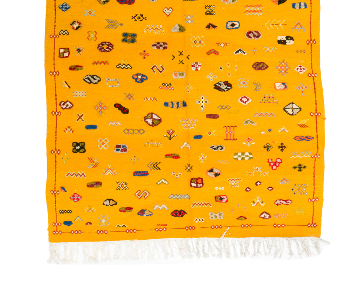 A yellow Moroccan rug with white fringe at the bottom and an all-over pattern of small multicolored geometric and tribal motifs.