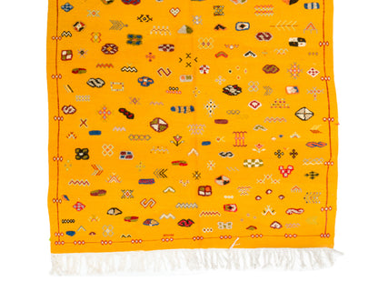 A yellow Moroccan rug with white fringe at the bottom and an all-over pattern of small multicolored geometric and tribal motifs.