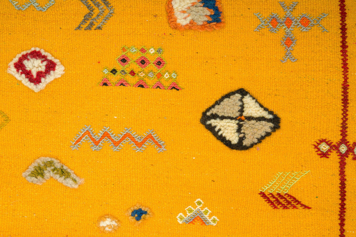 Close-up of a bright yellow woven rug with embroidered geometric motifs in red, blue, green, black, and white.