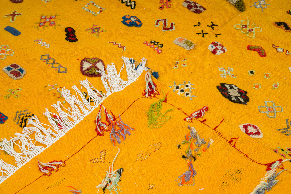 A close-up of a bright yellow Moroccan rug with white fringe, featuring multicolored geometric and floral woven patterns.