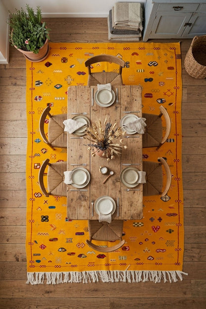 A top-down view of a bright yellow Moroccan rug with white fringe, styled with a wooden dining table and chairs on a hardwood floor.