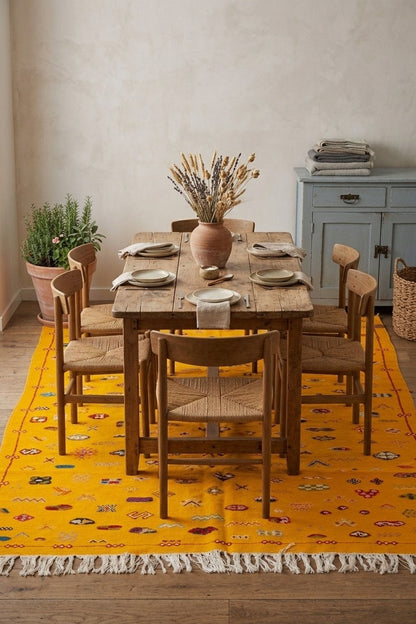 A rustic wooden dining table set with six woven-back chairs, styled on a bright yellow patterned area rug in a warm, neutral-toned dining room.
