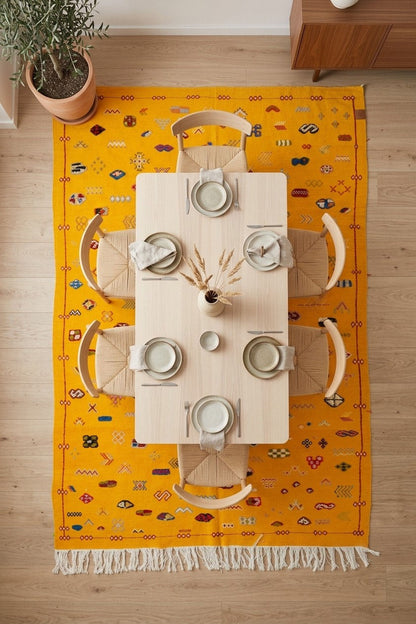A top-down view of a bright yellow Moroccan area rug with white fringe, styled with a light wood dining table and four chairs on a light wood floor.