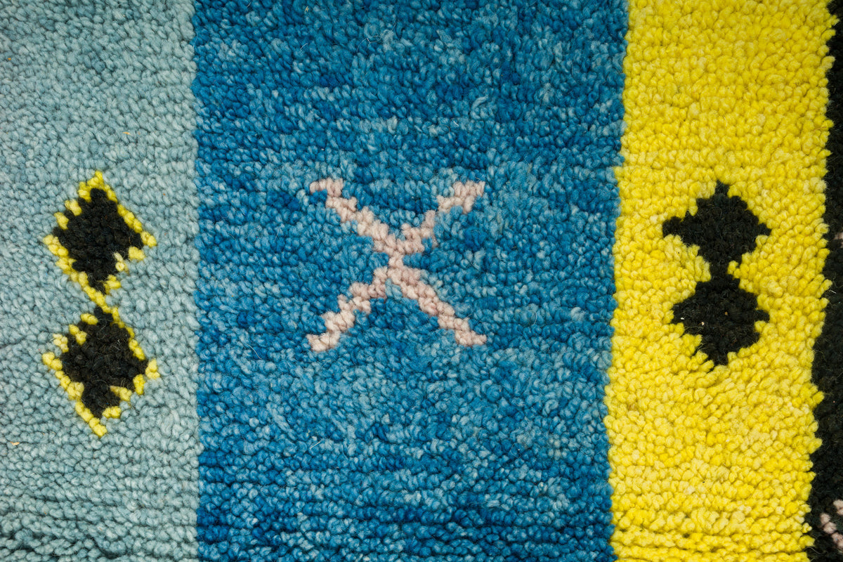 Close-up of a textured wool moroccan rug with vertical color blocks: light blue, medium blue, yellow, and black, with small geometric motifs including a pale pink X in the center blue section.