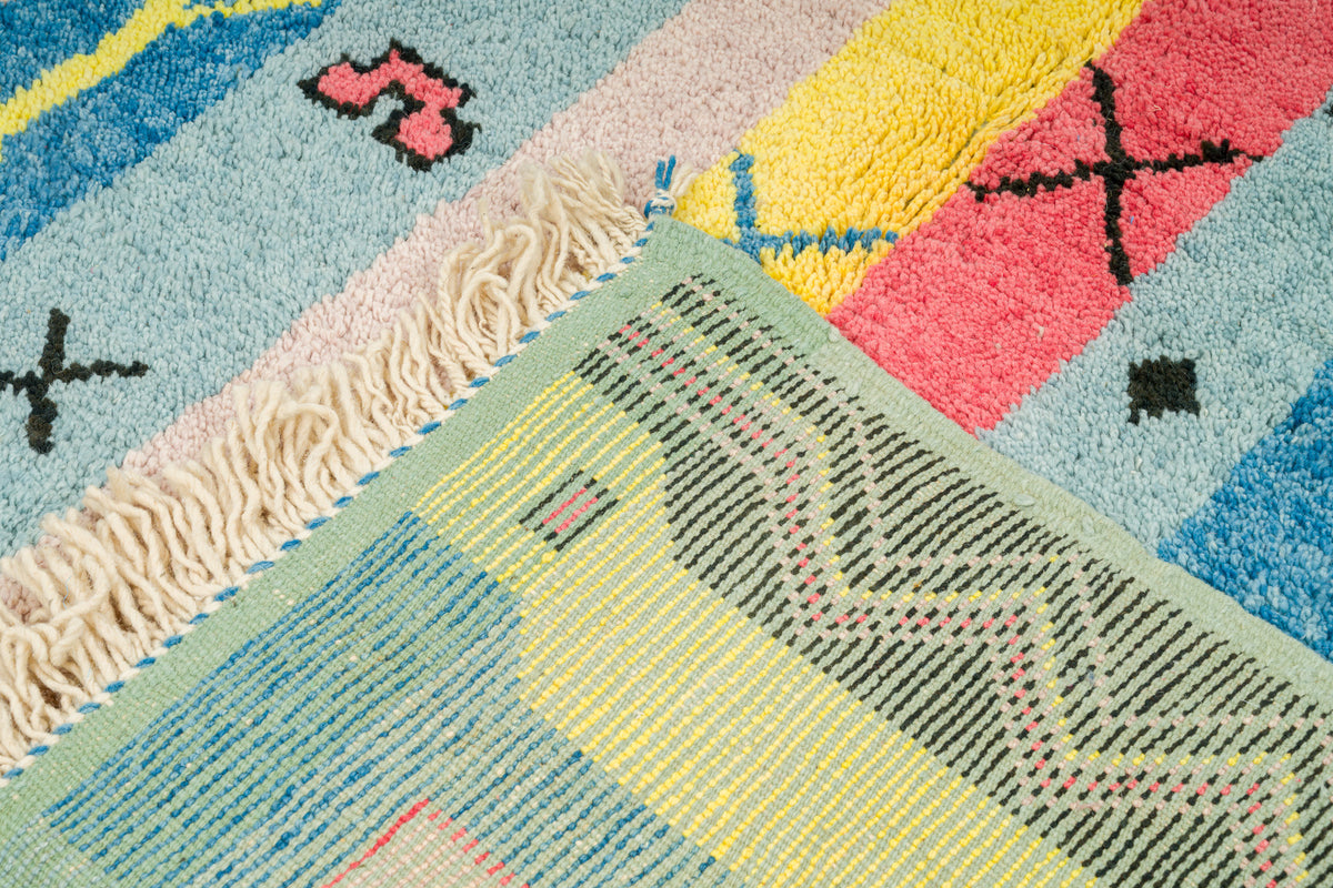 Close-up of a colorful hand knotted moroccan rug with geometric patterns, cream fringe, and a mix of pastel blue, yellow, pink, green, and black sections.