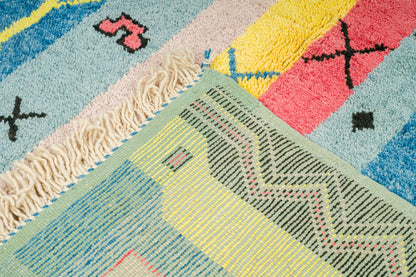 Close-up of a colorful hand knotted moroccan rug with geometric patterns, cream fringe, and a mix of pastel blue, yellow, pink, green, and black sections.