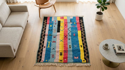 A colorful rectangular moroccan rug with vertical stripes and geometric motifs in blue, pink, yellow, black, and cream, styled on a light wood floor in a modern living room setting.