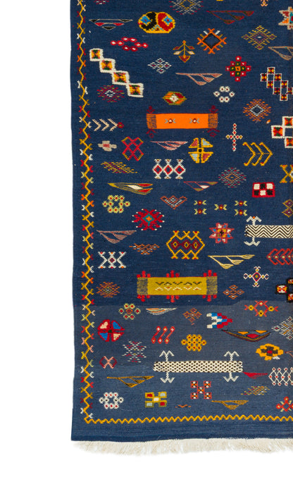 A close-up of a blue patterned rug with geometric and tribal-inspired motifs in red, yellow, white, and orange, featuring a fringed edge.