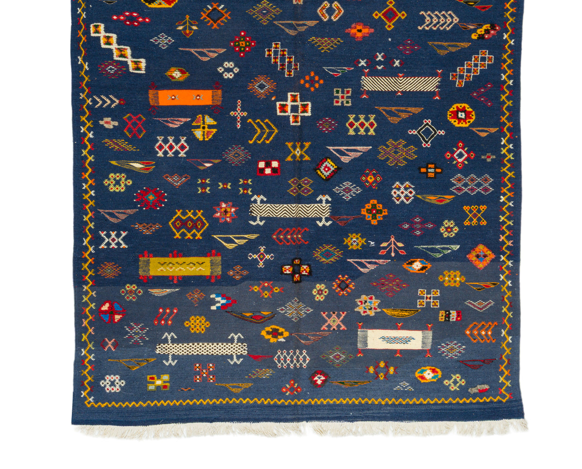 A dark blue wool rug with intricate multicolor geometric patterns and white fringe along the bottom edge, shown flat against a white background.