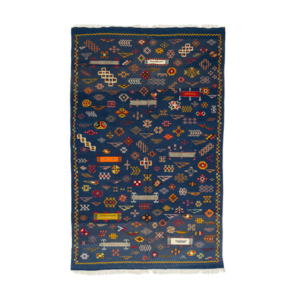 A rectangular dark blue wool rug with intricate multicolor geometric patterns and white fringe on the top and bottom, shown flat against a white background. The beautiful rug is moroccan.
