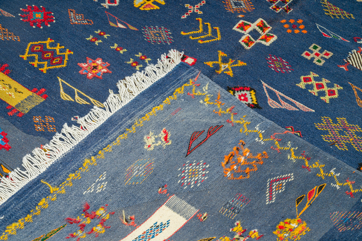 A close-up of a blue patterned rug with white fringe, showing geometric and floral motifs in red, yellow, orange, and white.