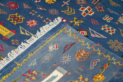 A close-up of a blue patterned rug with white fringe, showing geometric and floral motifs in red, yellow, orange, and white.