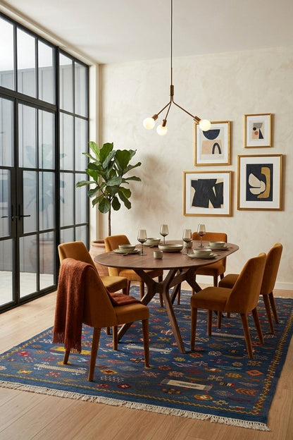 A styled dining room scene featuring a round wooden dining table surrounded by six mustard yellow upholstered chairs with wooden legs, set on a blue patterned area rug. A brown throw blanket is draped over one chair.