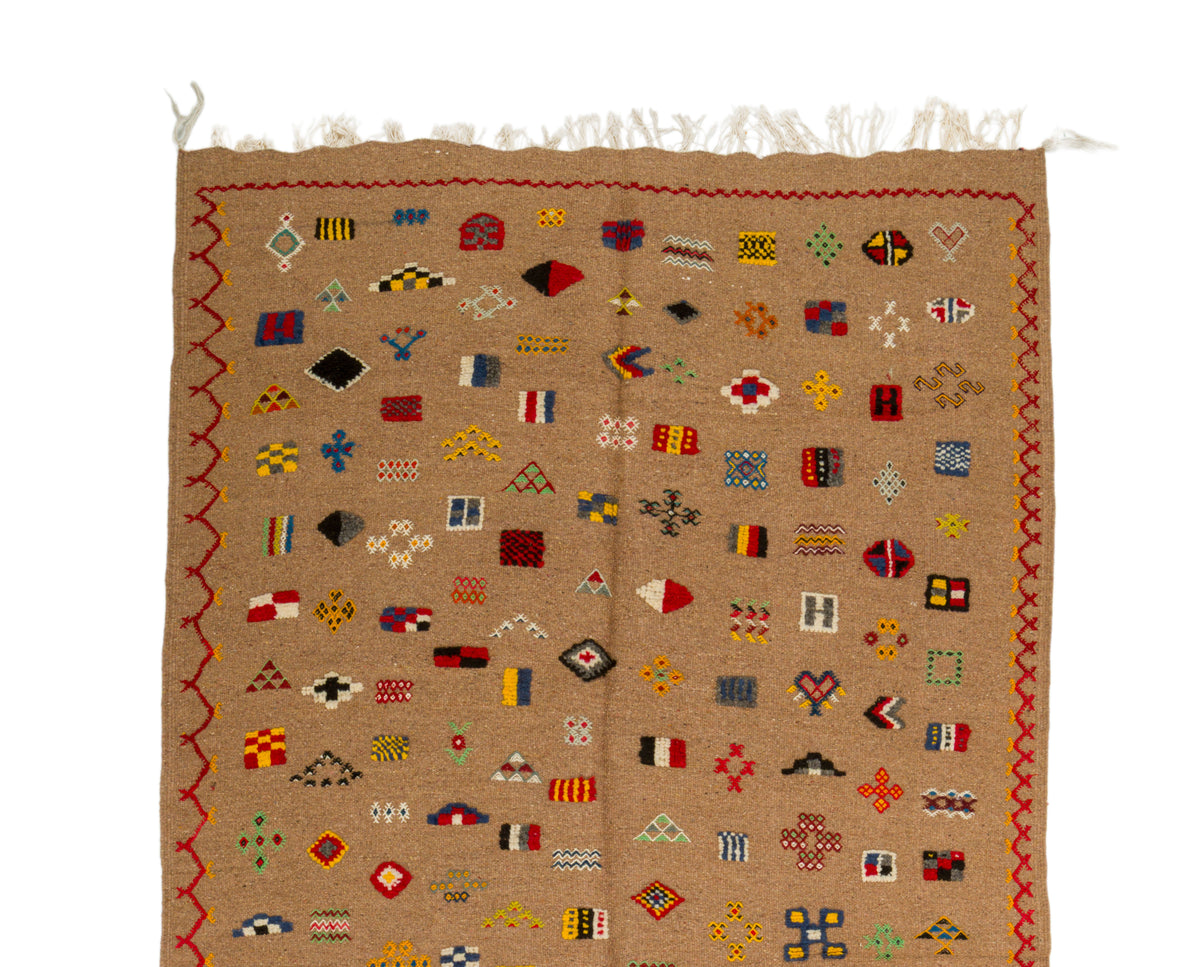 A square woven textile rug in tan with a multicolor geometric pattern of small flags and borders, shown flat against a white background.