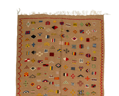 A square woven textile rug in tan with a multicolor geometric pattern of small flags and borders, shown flat against a white background.