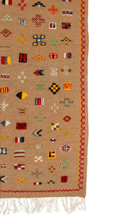 A close-up of a woven rug with a tan background, featuring a colorful geometric pattern and a red zigzag border, with white fringe at the bottom.