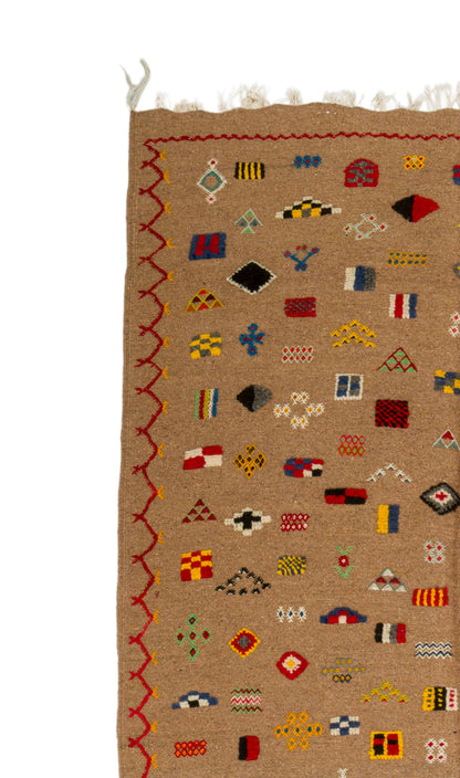 A close-up of a woven rug with a tan background, featuring a colorful geometric pattern of small flags and symbols, a red zigzag border, and white fringe at the top.