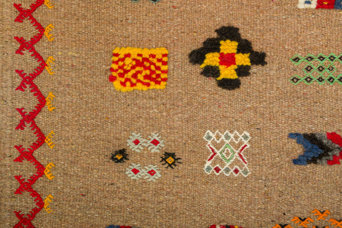 Close-up of a woven rug in a natural tan color, decorated with geometric and zigzag patterns in red, yellow, black, green, blue, and white.