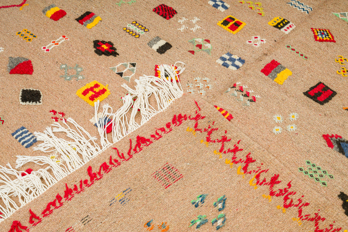A close-up of a woven rug with a beige base, featuring colorful geometric motifs in red, yellow, black, blue, and white, along with white fringe on one end.