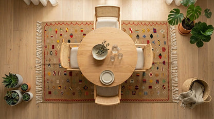 A top-down view of a round wooden dining table surrounded by four woven chairs on a patterned beige rug with fringe, placed on a light wood floor. Small potted plants and a basket with a throw blanket are arranged around the scene.