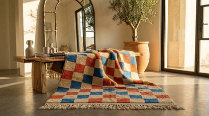 A multicolor checkered wool rug with fringe draped over a rustic wooden bench in a sunlit, modern interior room with a potted olive tree. The rug is a Moroccan rug.