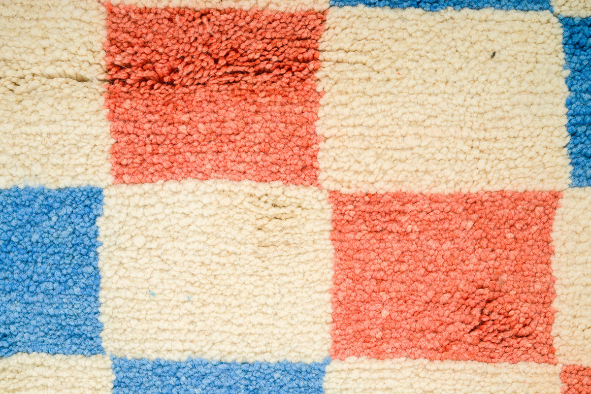 A close-up of a textured rug with a checkered pattern in cream, coral red, and blue.