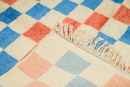 A close-up of a Moroccan woven rug with a checkered pattern in cream, blue, and coral, featuring fringe along one edge.