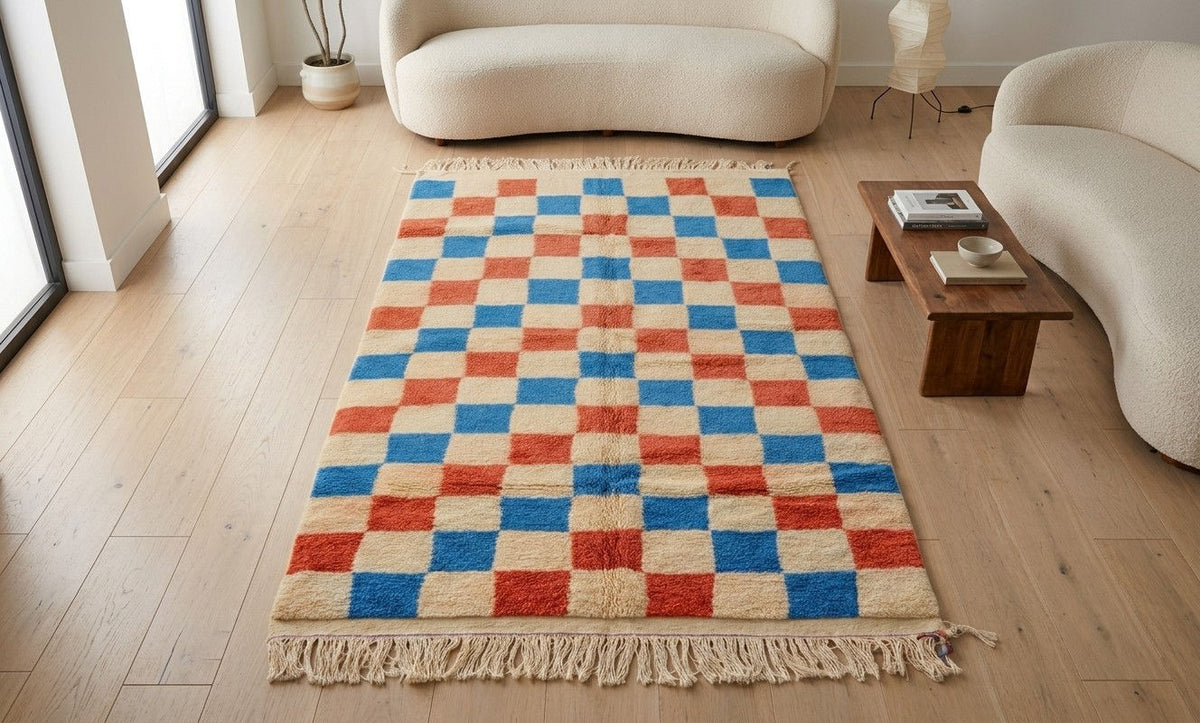 A Moroccan high pile woven area rug with a checkered pattern in red, blue, and cream, featuring fringe on the ends, displayed on a light wood floor in a modern living room setting.