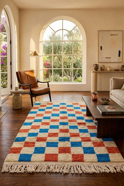 A rectangular area rug with a cream base and a bold checkerboard pattern in blue and terracotta red, styled in a bright living room with a wooden coffee table, leather armchair, and large arched windows. A moroccan rug.