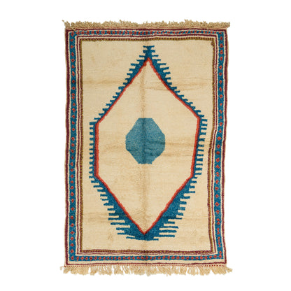 A rectangular wool hand-woven rug with a cream-colored base, featuring a central blue geometric diamond pattern outlined in red, surrounded by a patterned border and fringed edges. The rug is a moroccan rug.