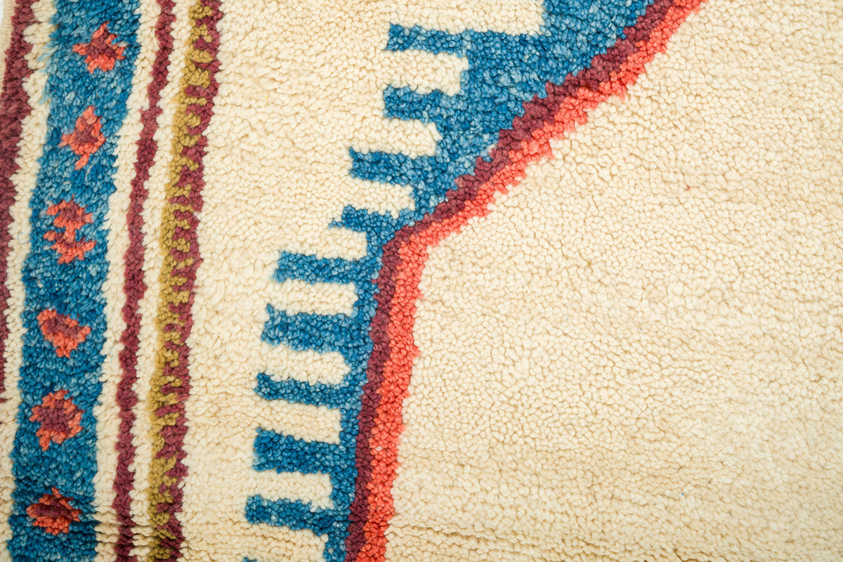 Close-up of a textured wool rug with a cream base, featuring a bold blue stepped pattern outlined in red and vertical striped bands with red floral motifs.