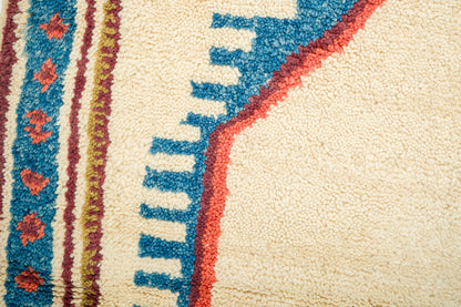 Close-up of a textured wool rug with a cream base, featuring a bold blue stepped pattern outlined in red and vertical striped bands with red floral motifs.