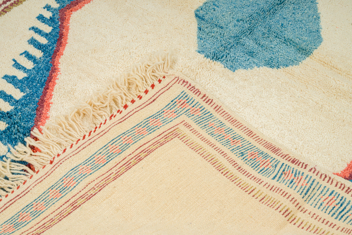 Close-up of a woven rug corner showing cream base, blue geometric shapes, and red and blue patterned fringe borders.