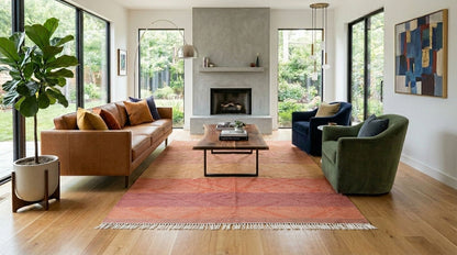 A modern living room with a large rust-red patterned Zanafi area rug on a light wood floor, featuring a brown leather sofa, a green armchair, a dark blue accent chair, and a central fireplace.
