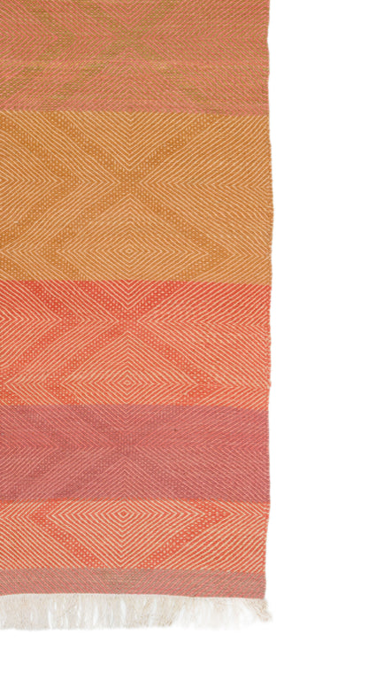 A close-up of a Moroccan Rug with horizontal bands of terracotta, mustard, coral, and dusty rose, featuring geometric diamond patterns and white fringe at the bottom, shown against a white background.